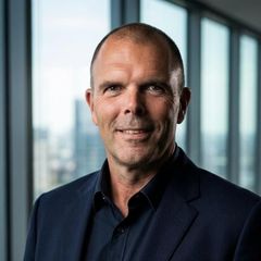 Man in dark suit smiles, posed in front of cityscape windows.