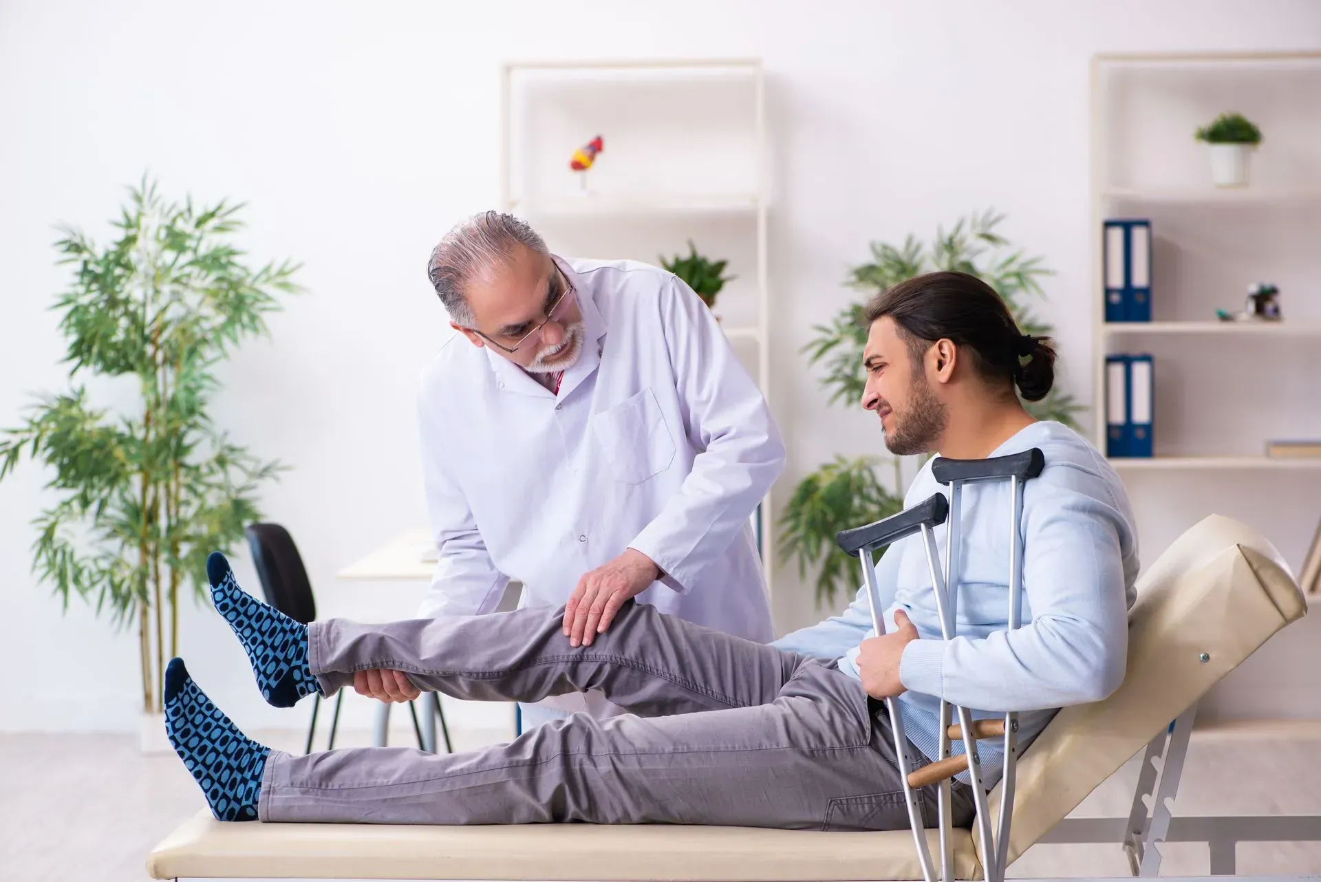 Doctor examines a man's injured knee; man on a medical exam table with crutches, indoor setting.