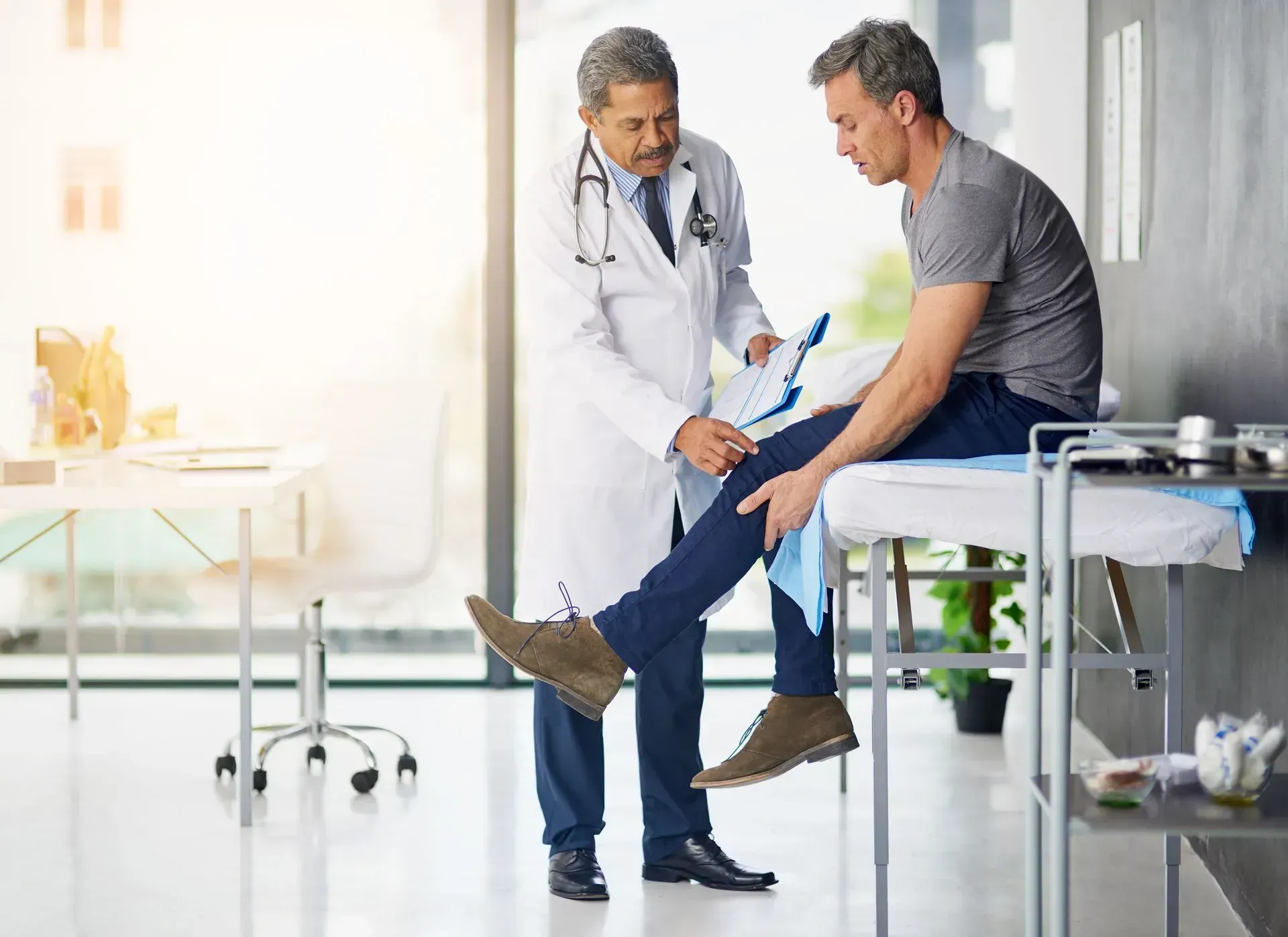 Doctor examining patient's knee in a clinic. Doctor in white coat holds clipboard, patient sits on examination table.