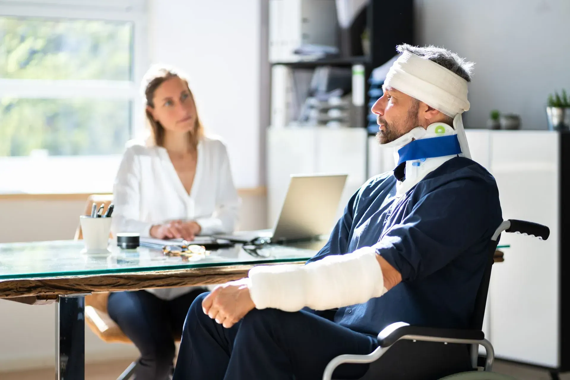 Man in wheelchair with bandages, neck brace, and cast, at a table with a woman in an office.