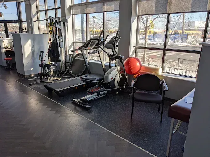 Gym interior with exercise equipment near a large window. Treadmill, elliptical, ball, and chairs present.