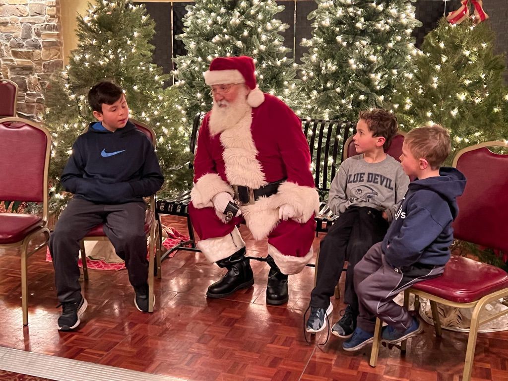 Santa Claus with three children, surrounded by Christmas trees, seated on chairs, talking.