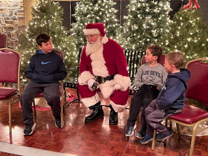 Santa Claus with three children, surrounded by Christmas trees, seated on chairs, talking.