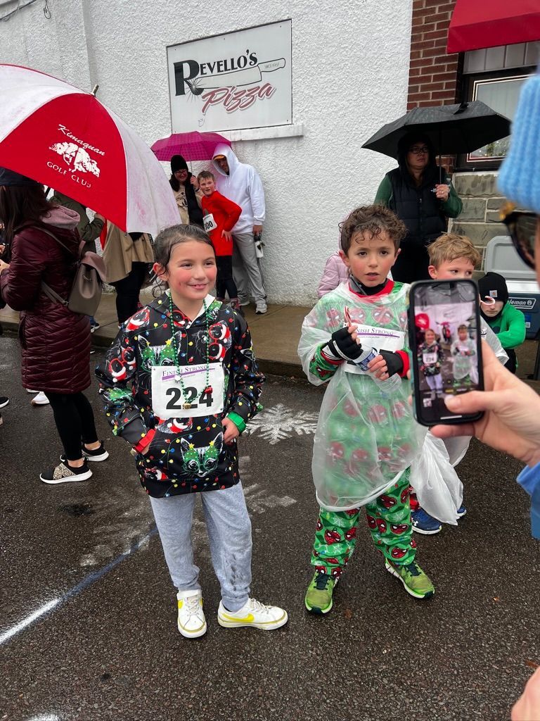 Two children in holiday outfits at an outdoor event, one holding a toy. It's raining, and people hold umbrellas.
