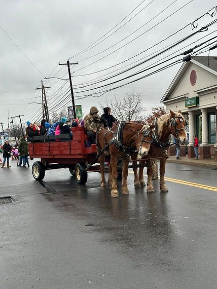 Horse-drawn wagon with people moves down street. Brown horses, red wagon, cloudy day.