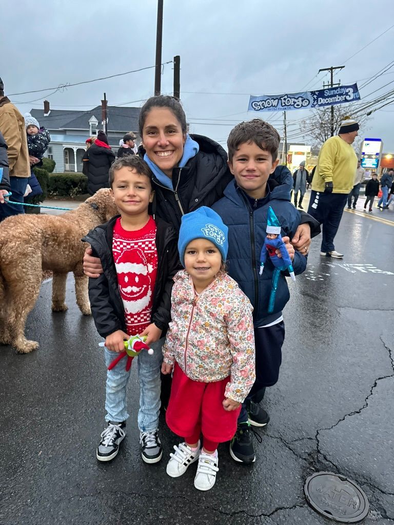 Woman and three children pose on a wet street. Boy in Santa sweater, another in blue jacket holds a toy.