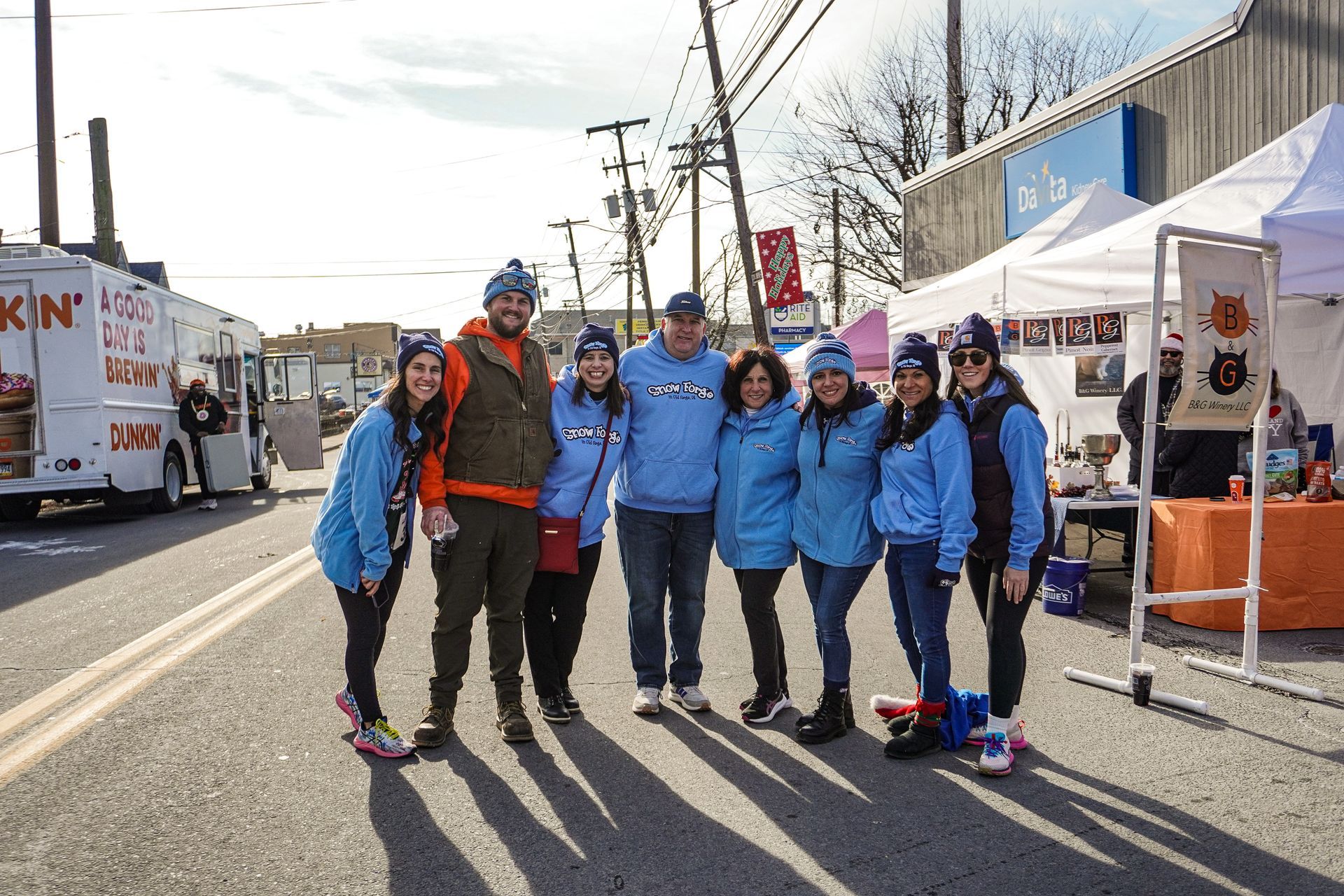 Group of people smiling, wearing matching light blue jackets and hats, posing outdoors in front of a food truck and tents.