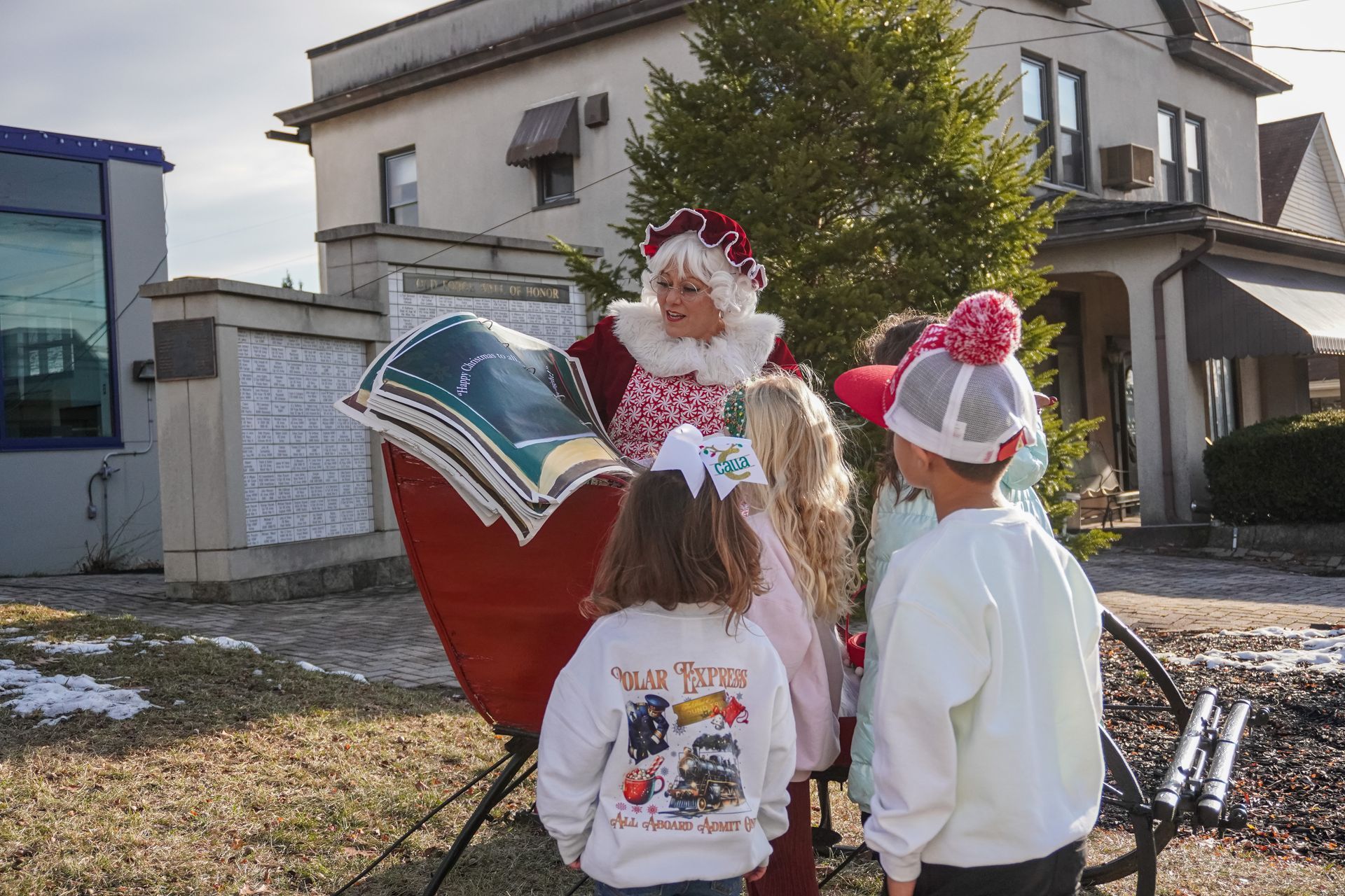 Mrs. Claus reading to children from a sleigh-shaped book in front of a house on a sunny day.