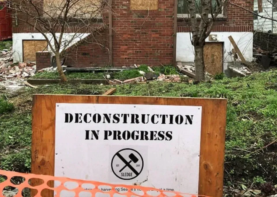 A sign that says deconstruction in progress is in front of a brick building.