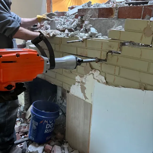 A man is using a demolition hammer to demolish a brick wall