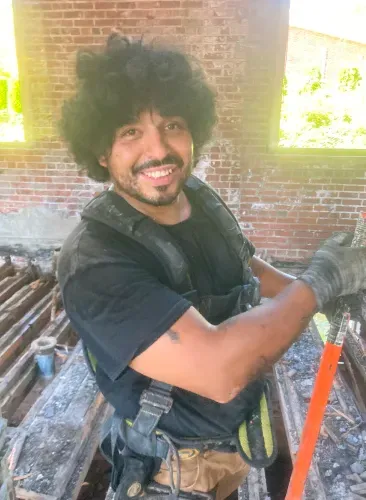 A man with curly hair and a beard is smiling while standing in front of a brick wall.