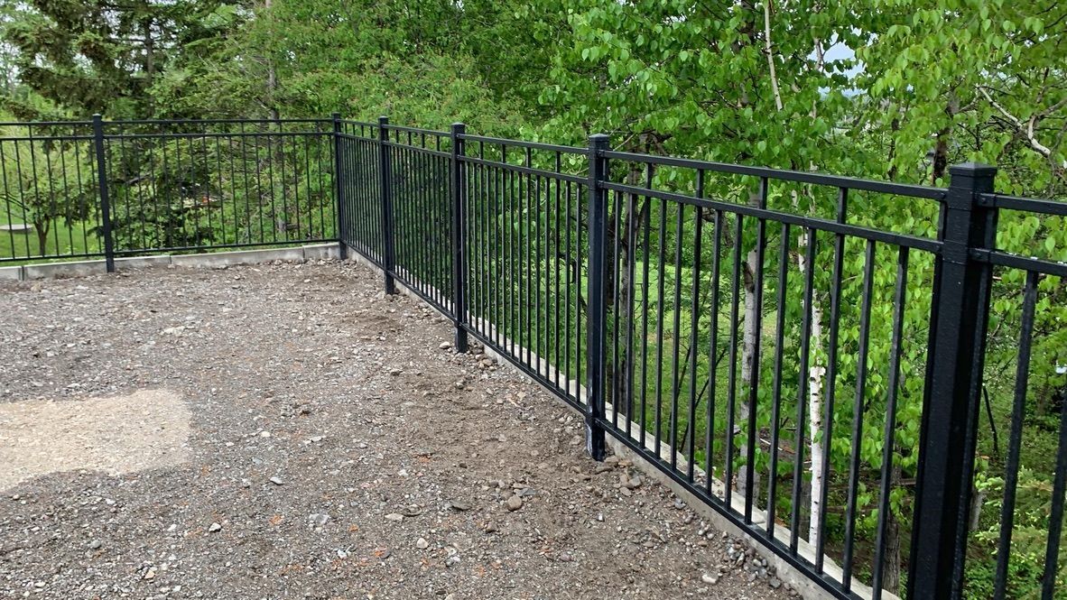 Une clôture en métal noir entoure une zone de gravier avec des arbres en arrière-plan.
