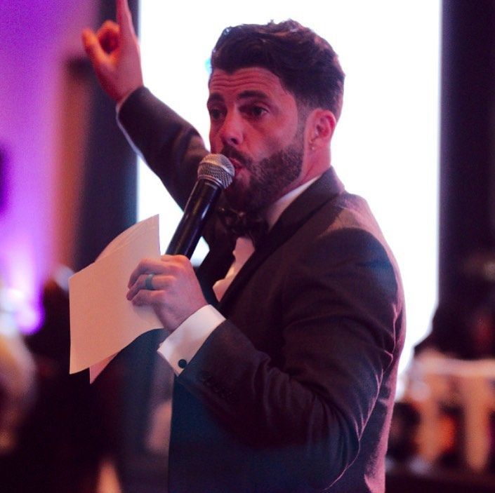 A man in a tuxedo is holding a microphone and giving a speech