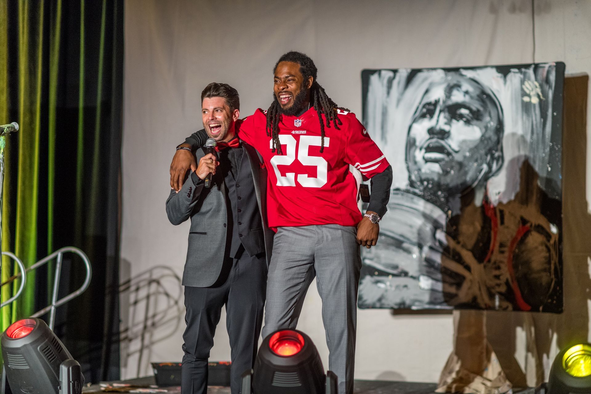 A man in a suit and a man in a red jersey are standing next to each other on a stage.