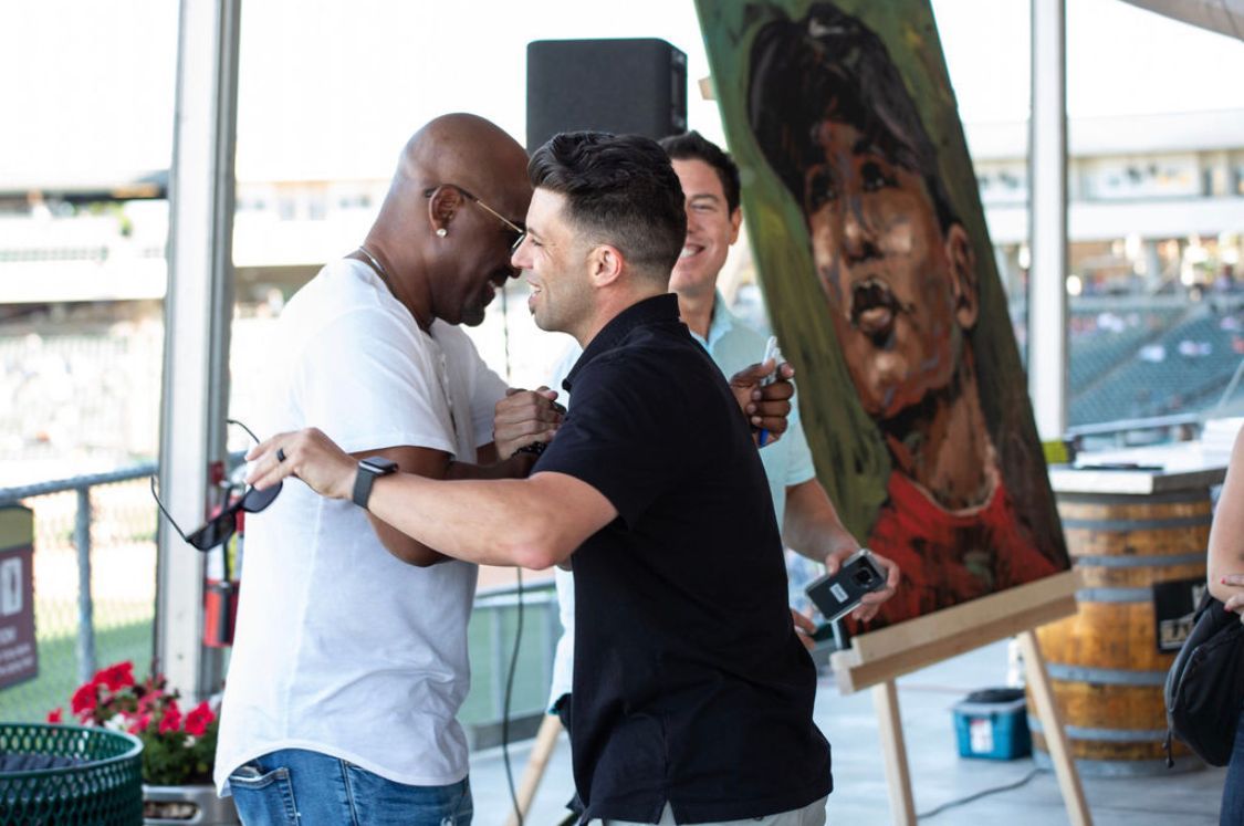 Two men are hugging in front of a painting of a baseball player.