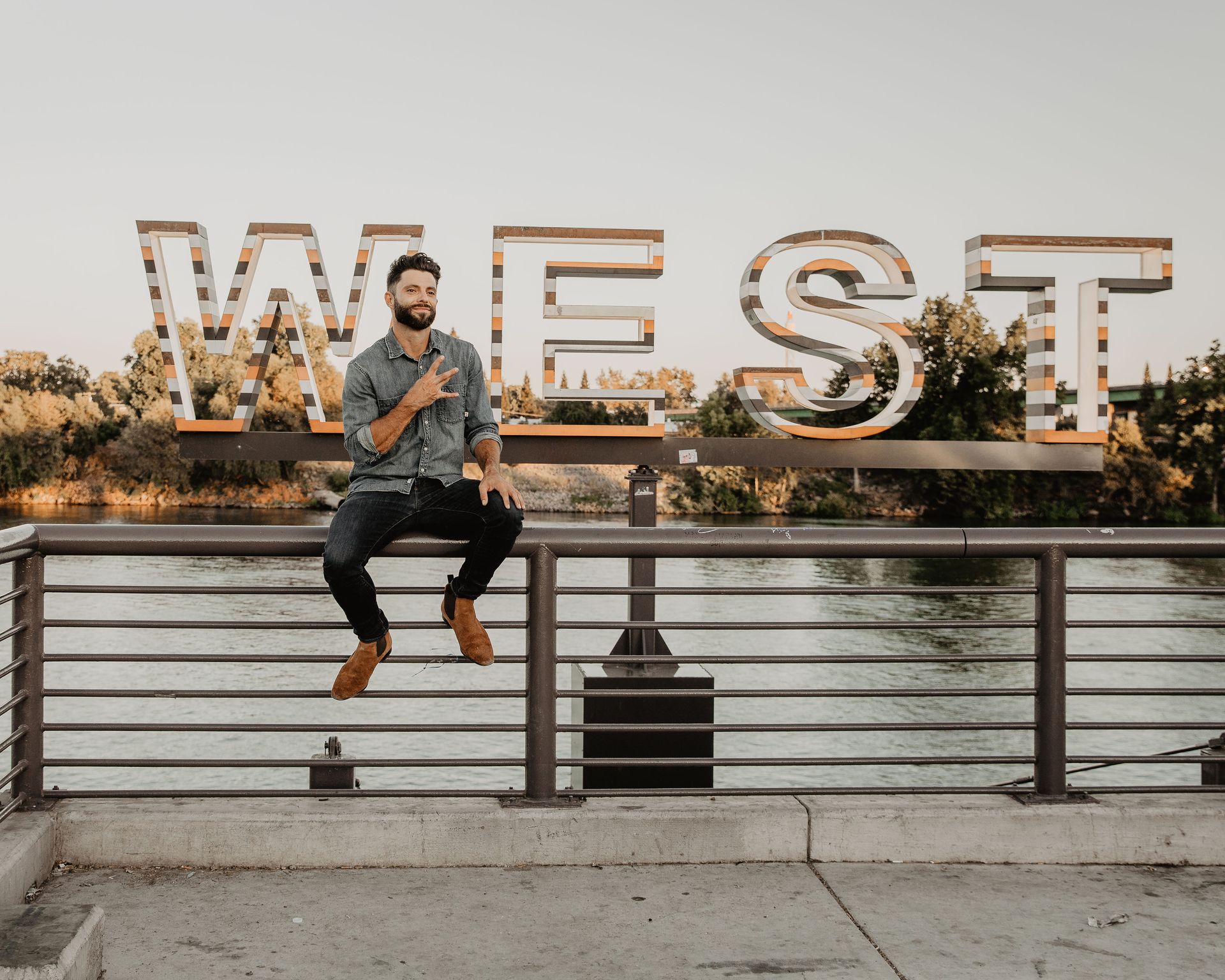 A man is sitting on a railing next to a sign that says west.