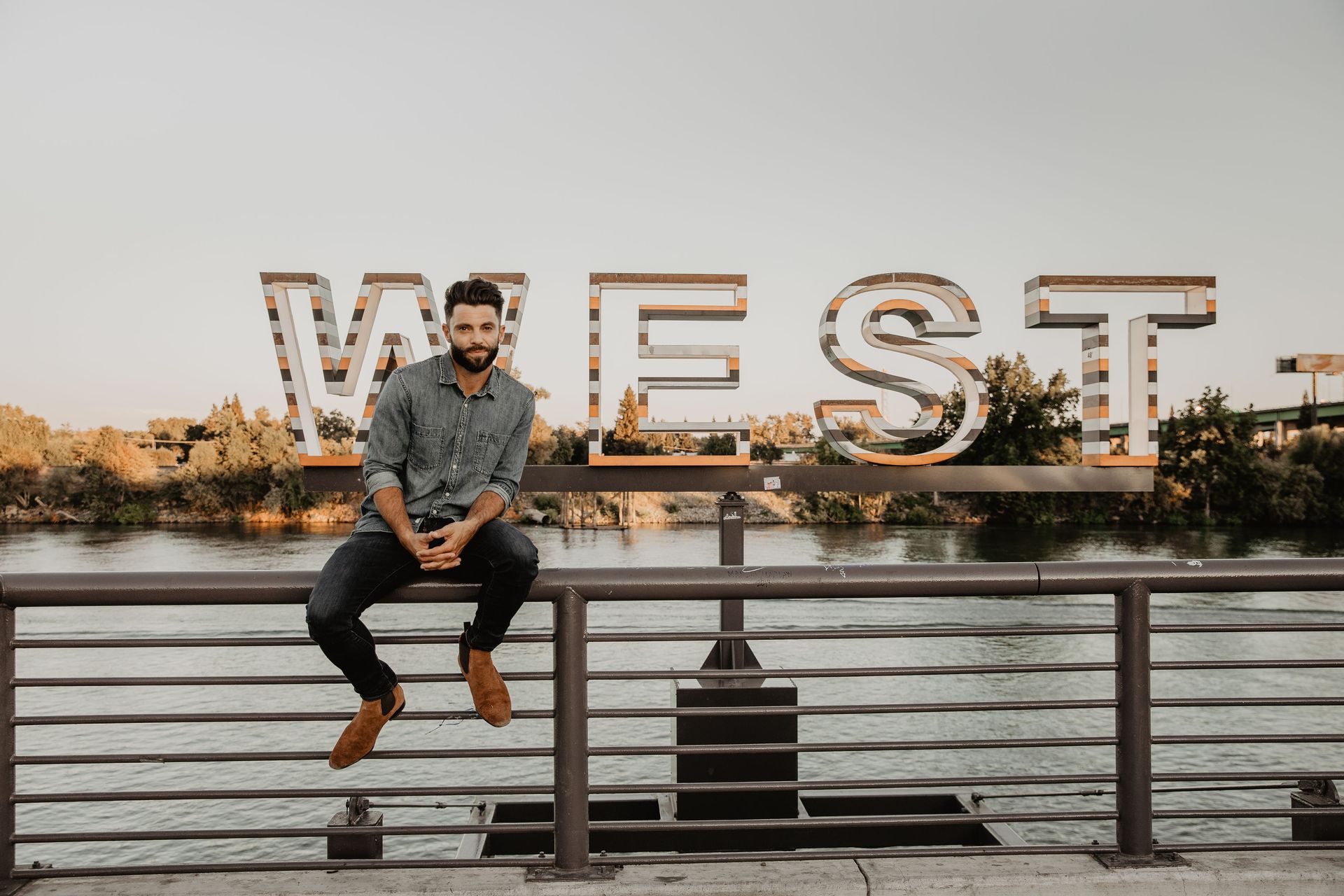 A man is sitting on a railing next to a sign that says west.