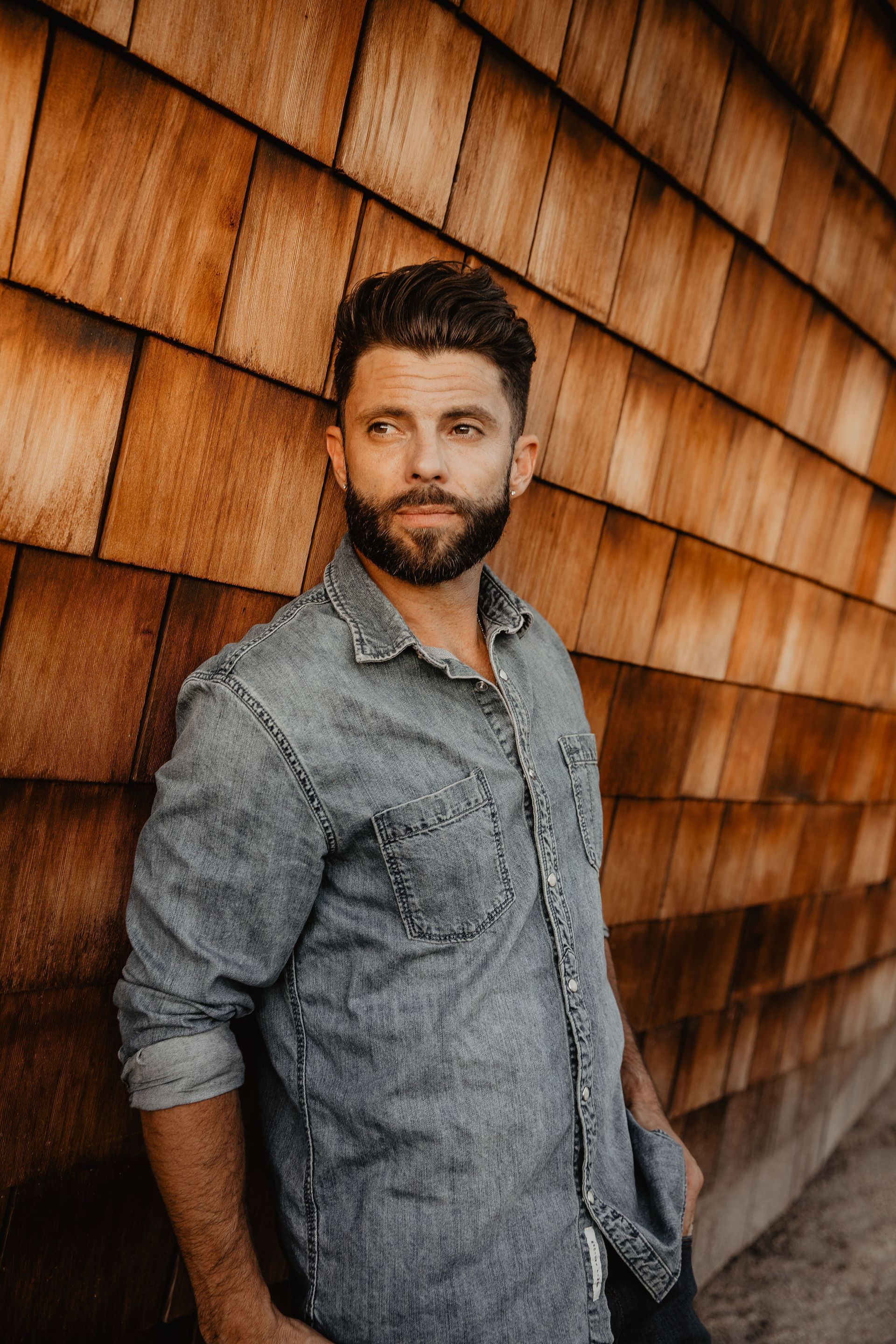 A man with a beard is leaning against a wooden wall.