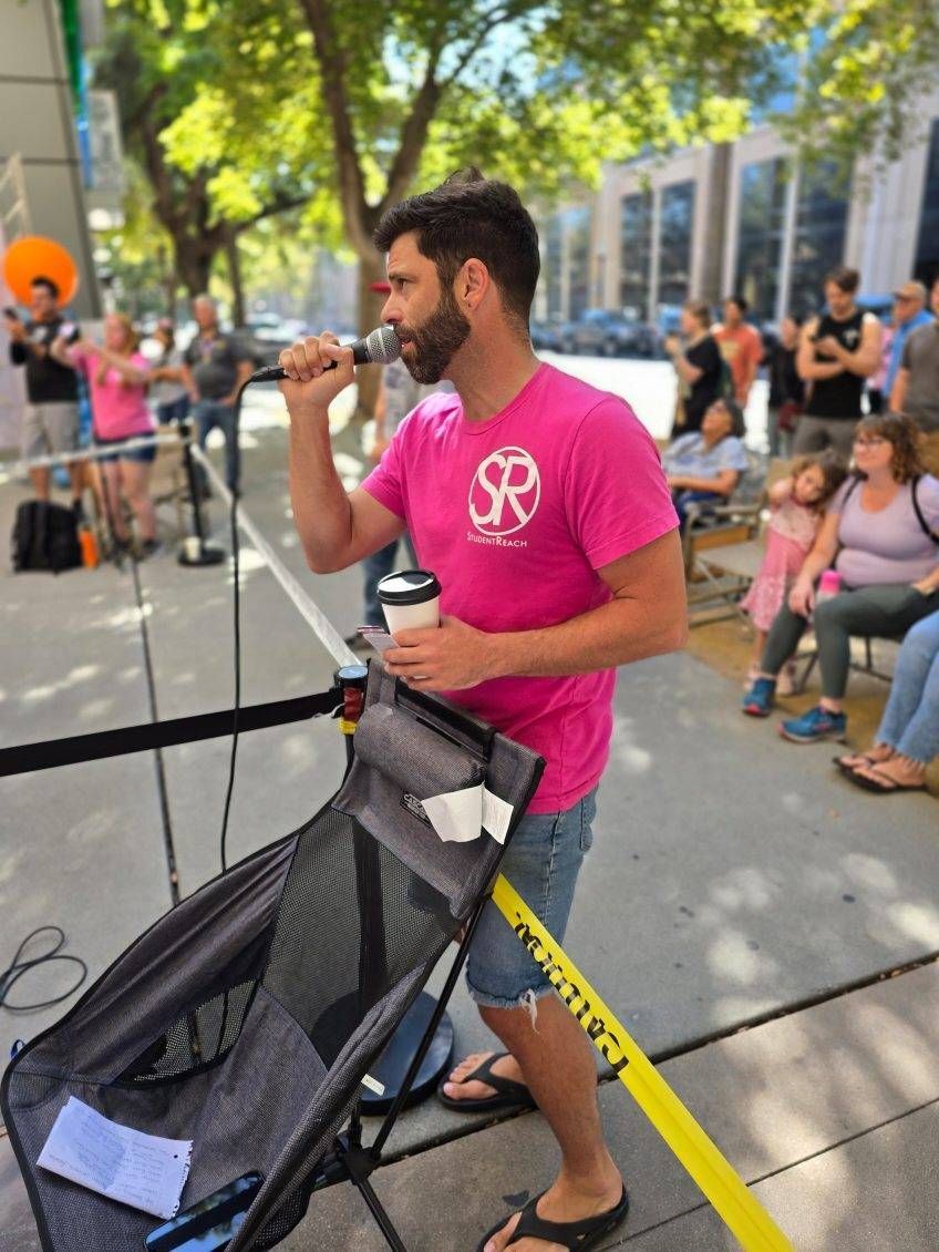 A man in a pink shirt is singing into a microphone while standing next to a stroller.