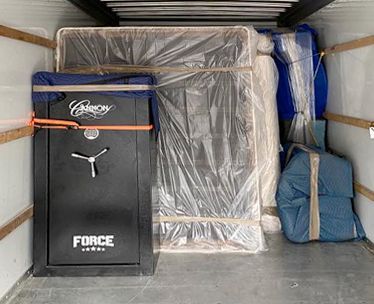 Inside a moving truck: a large black gun safe, wrapped furniture, and a mattress.