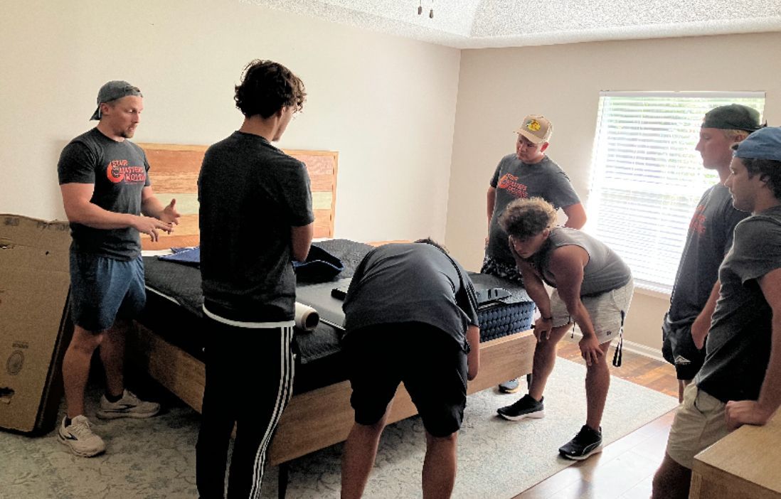 Men assembling a bed in a bedroom; some look at instructions, others handle parts.