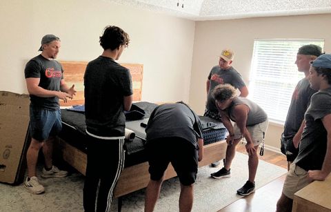 Men assembling a bed in a bedroom; some look at instructions, others handle parts.