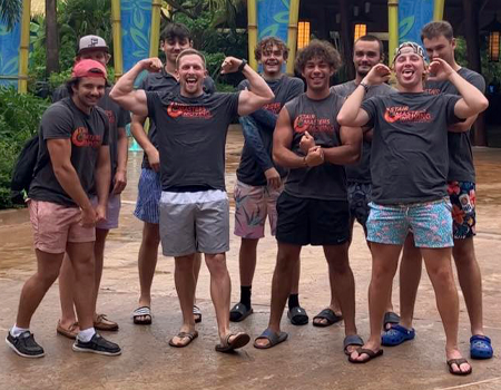 Group of men flexing at a water park. They wear t-shirts and swim trunks, smiling. Outdoors