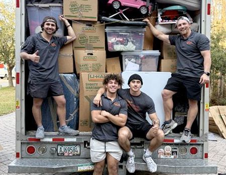 Four movers in gray shirts and shorts pose in front of a packed moving truck, boxes stacked high.
