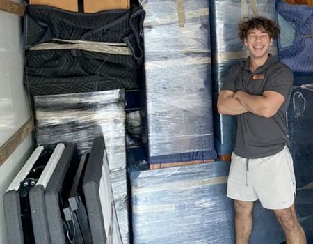 Man standing in a moving truck, smiling with arms crossed, surrounded by wrapped furniture.