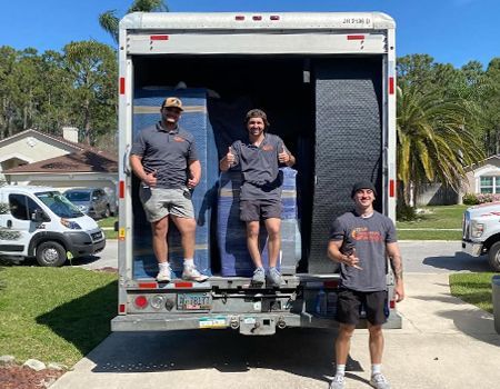 Three movers stand in the back of a moving truck, smiling. Two blue furniture pads and a wall are visible.