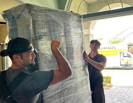 Two movers lift a large, wrapped object in a doorway. One flexes, the other assists. A U-Haul truck is visible.