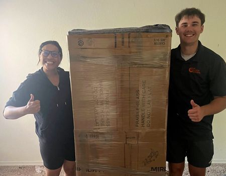 Two people smiling, giving thumbs up, beside a large wrapped cardboard box indoors.