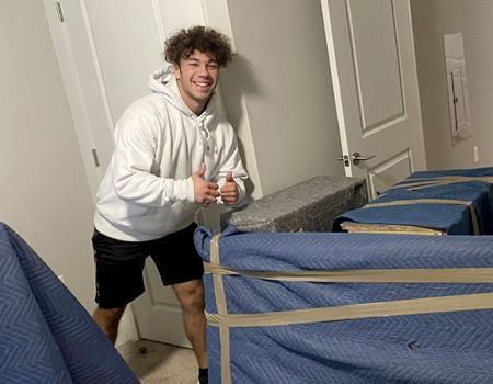 Young man smiling, giving thumbs up, near wrapped furniture in an empty room, possibly moving.