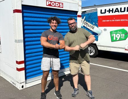 Two men flexing arms in front of a blue PODS storage unit and a U-Haul truck.
