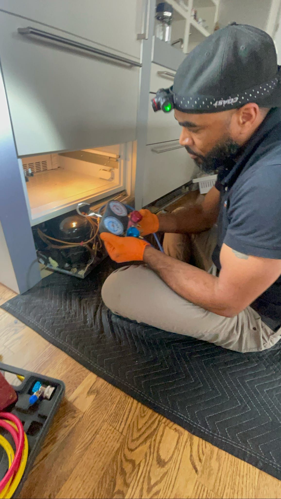 A man is kneeling on the floor fixing a refrigerator.