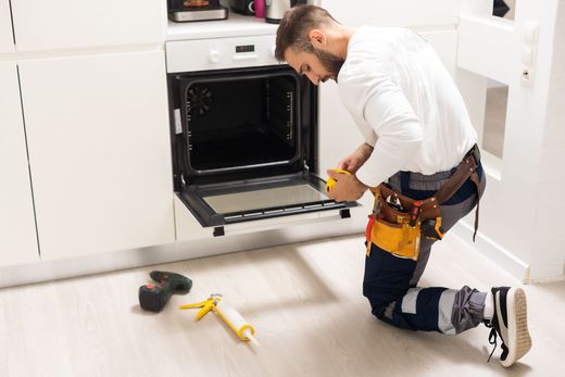 A man is kneeling down in a kitchen fixing an oven.