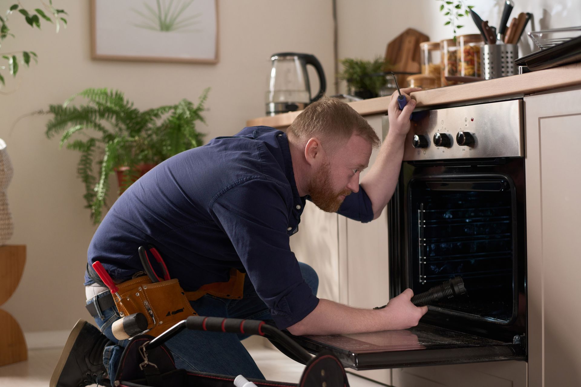 A man is kneeling down in a kitchen fixing an oven.