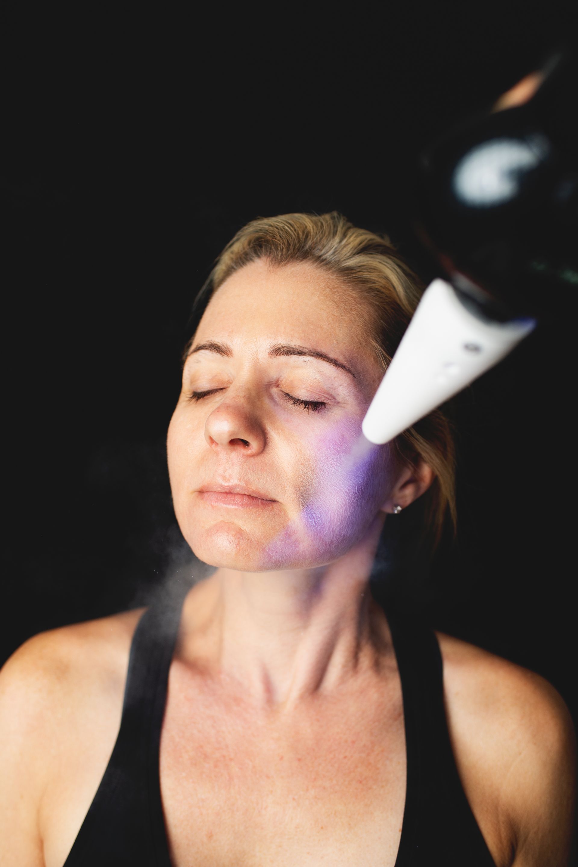 Woman receiving facial treatment, device emitting purple light and mist. Black background.