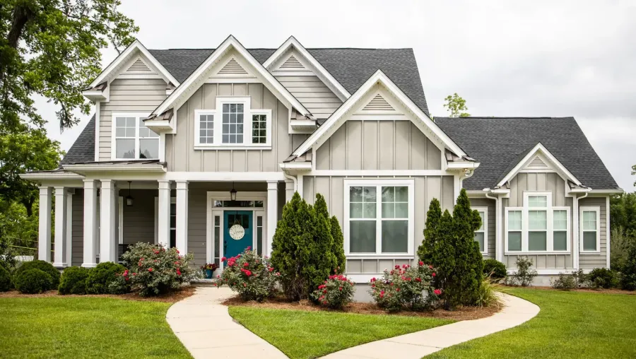 Large beige two-story suburban house with a manicured lawn, curved walkway, and flowering shrubs