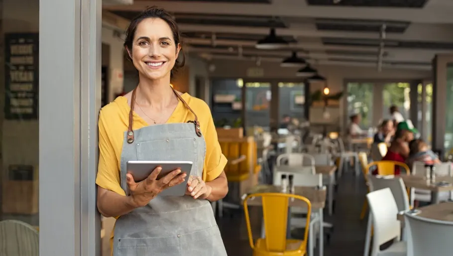 Smiling café worker in yellow shirt and gray apron holding a tablet inside a modern restaurant