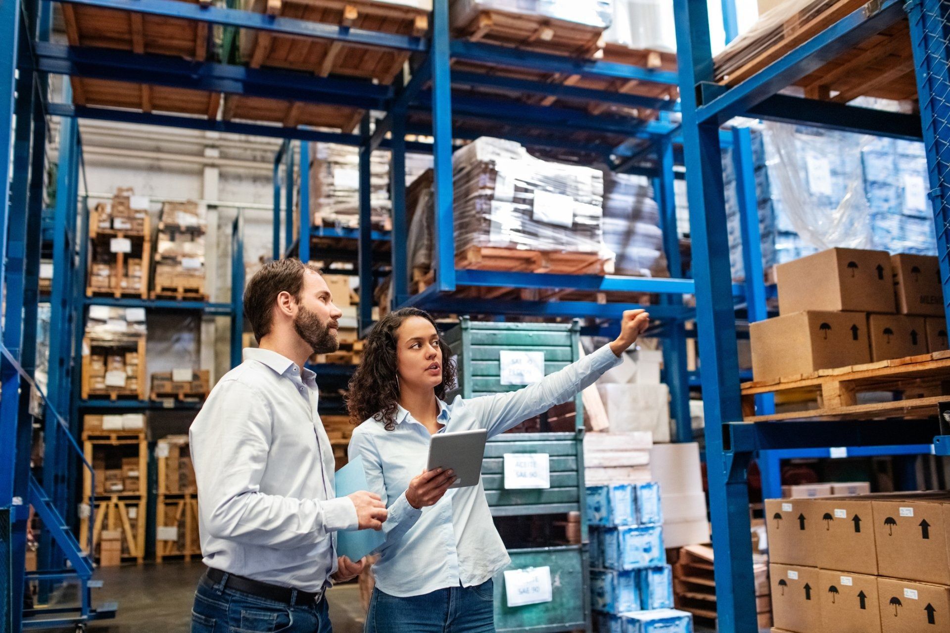 Two workers reviewing a clipboard in a warehouse with stacked boxes and pallets