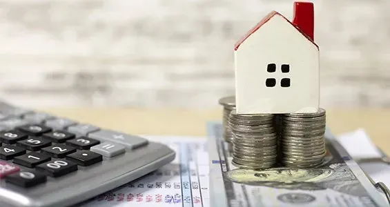 House model on stacked coins beside calculator and bills, symbolizing mortgage or home savings.