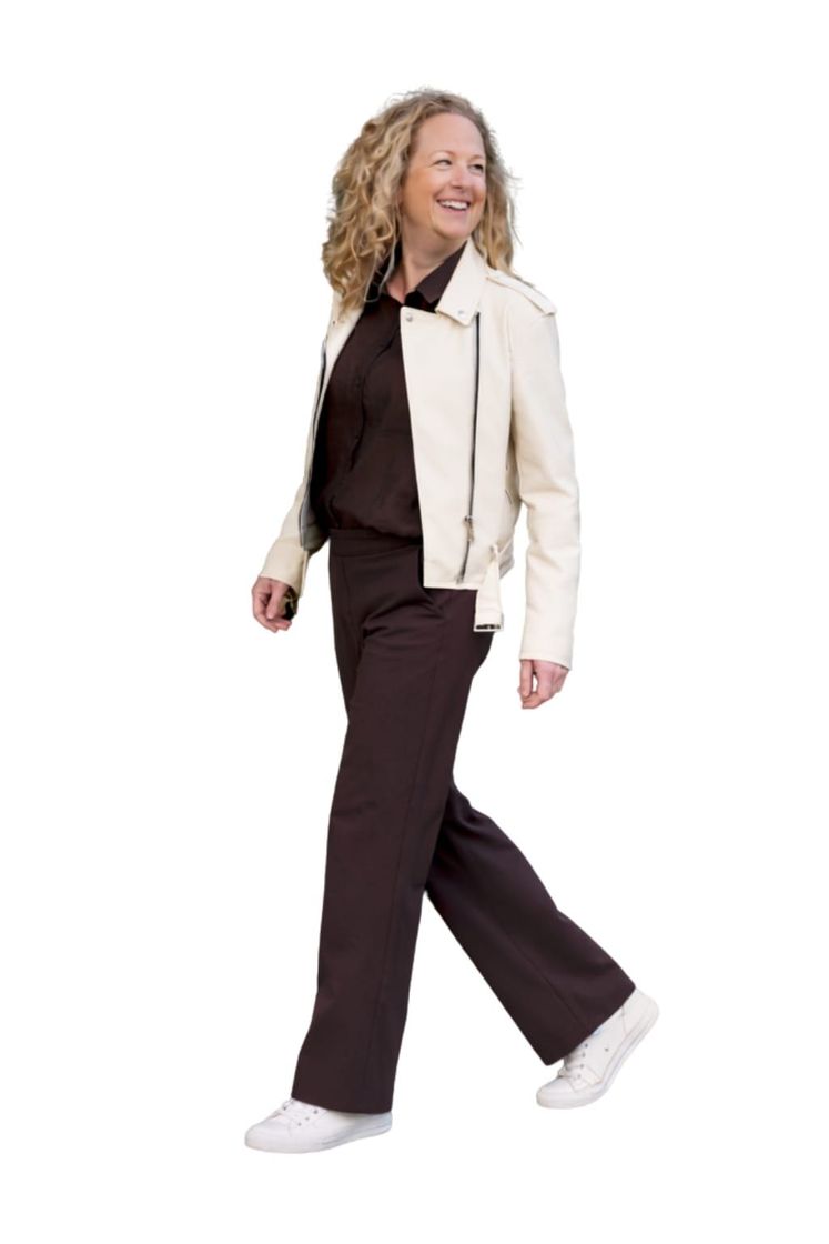 Een persoon met krullend haar die lachend loopt, gekleed in een crèmekleurig motorjack, een donkere broek en witte sneakers.