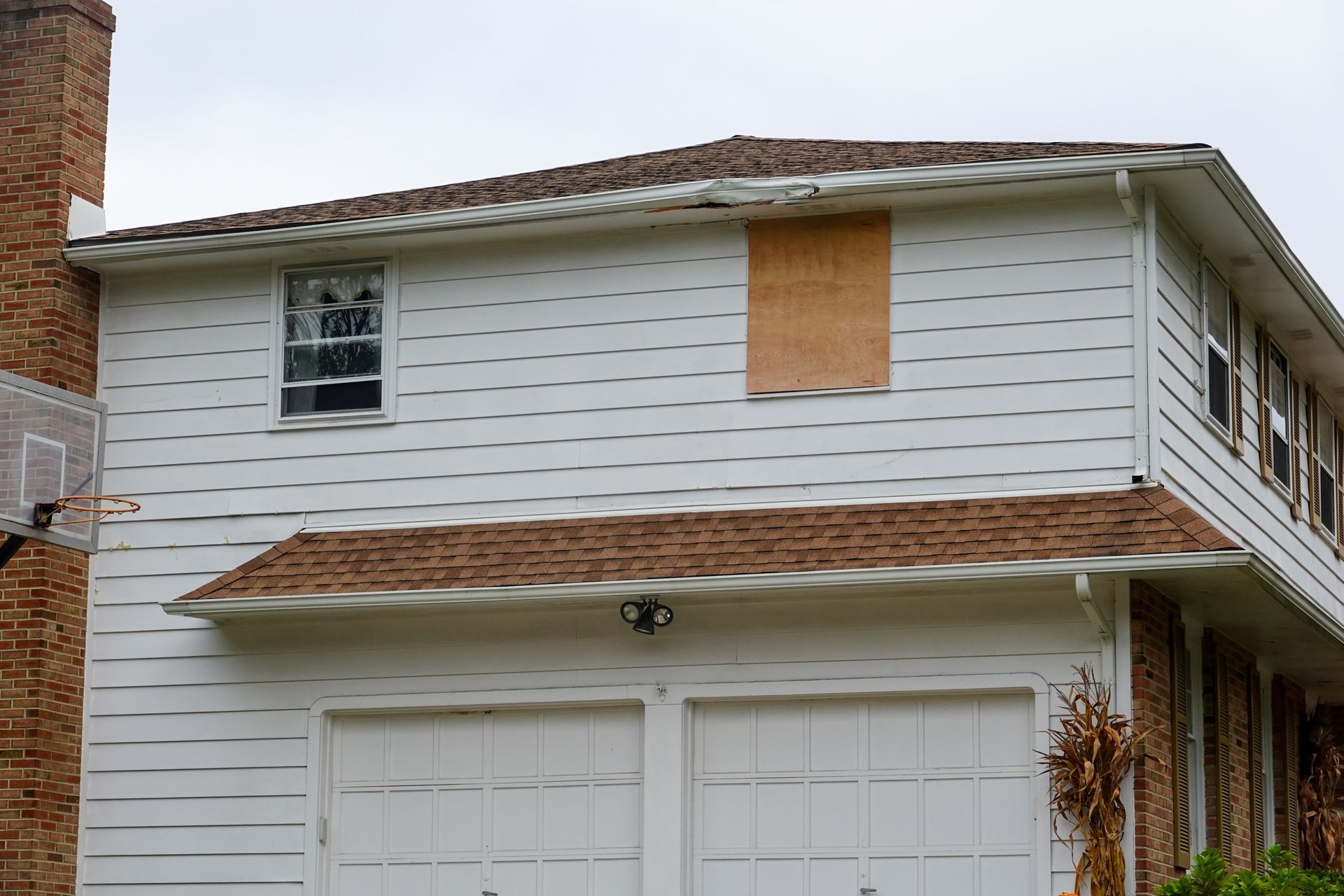 White two-story house with boarded-up window, brick chimney, and attached garage under a cloudy sky.