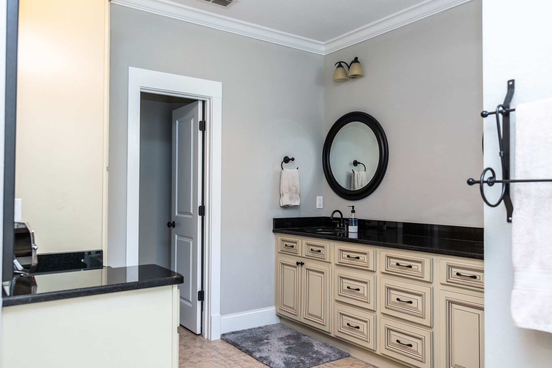 Bathroom with light gray walls, black countertop, cream cabinets, and a round mirror.