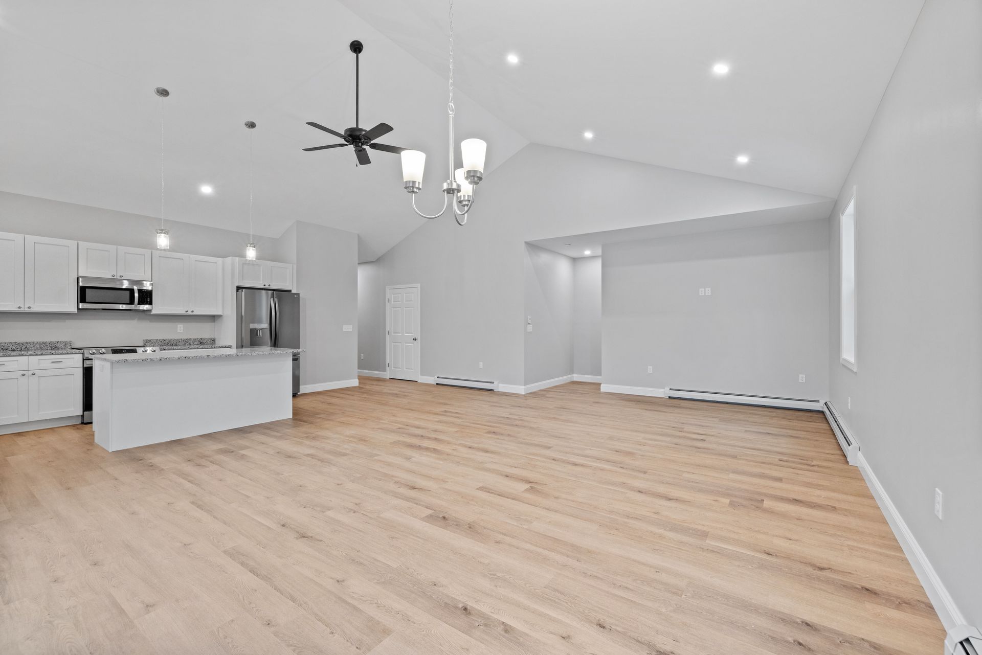 Empty, modern living room with light wood floors, gray walls, and open kitchen.