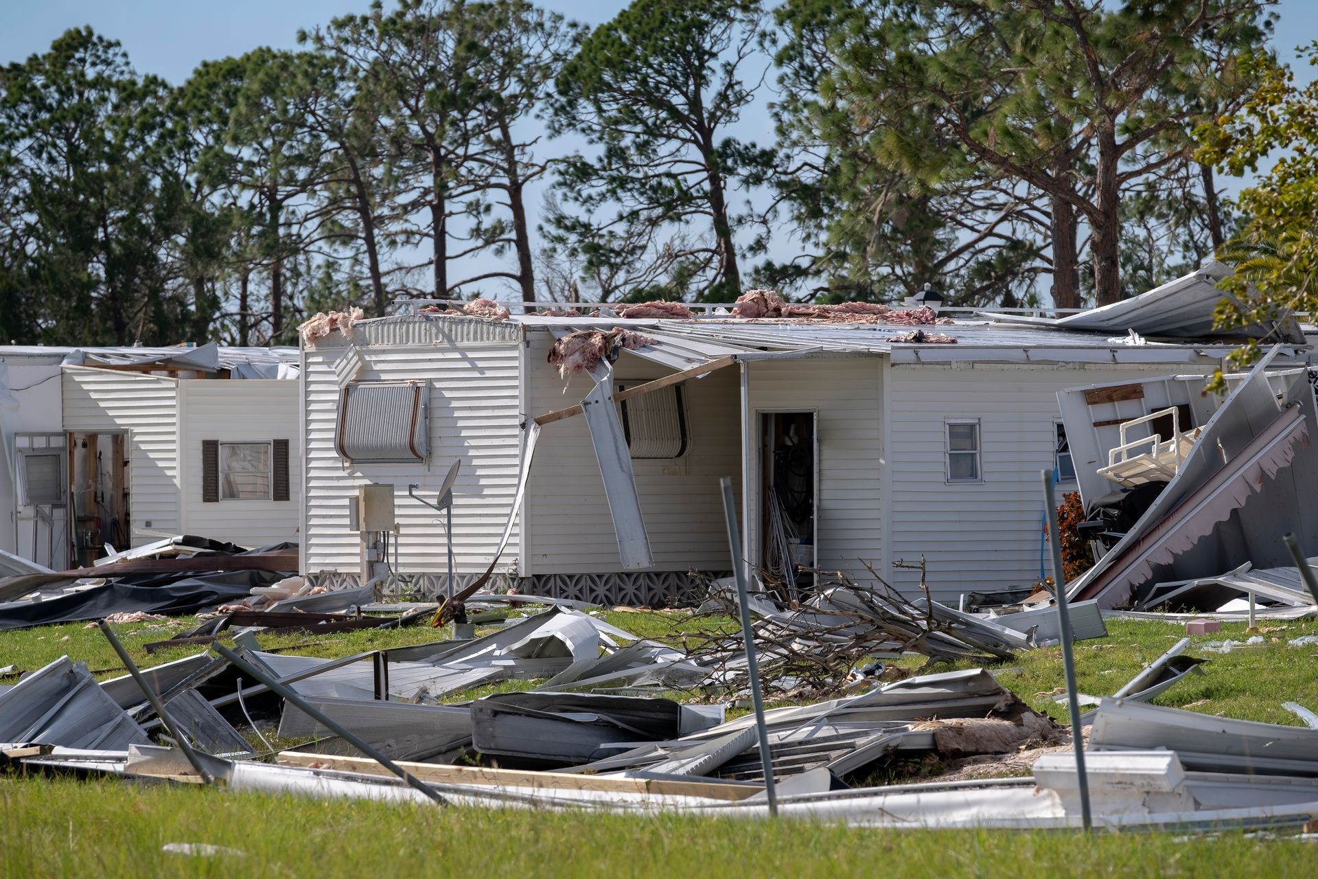 Damaged mobile homes with collapsed roofs and debris on the ground, trees in the background.