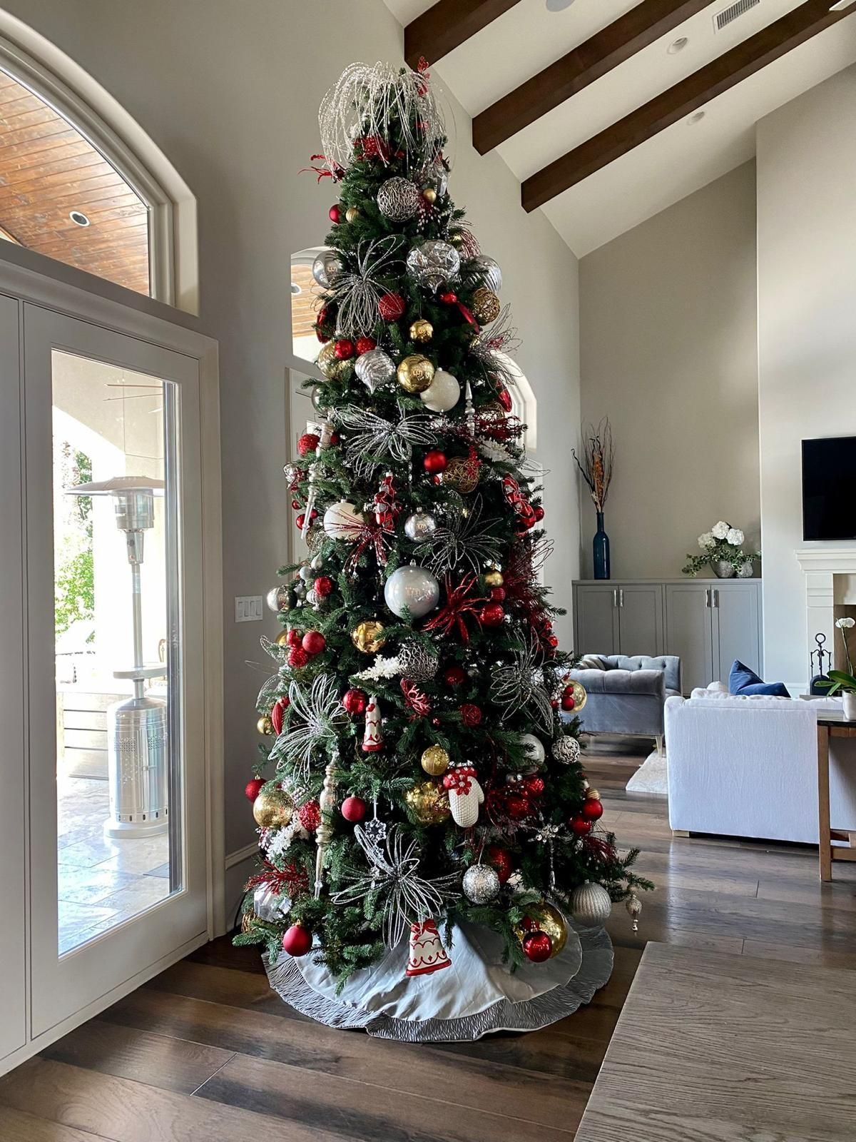 A tall, decorated Christmas tree with red, gold, and silver ornaments stands in a bright room with high, beamed ceilings.