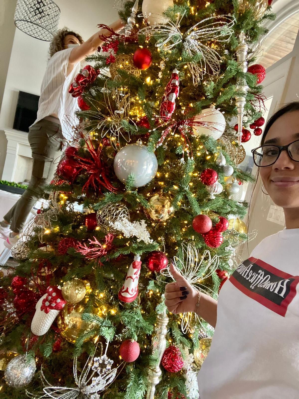 Two people decorate a large, lit Christmas tree with red, gold, and white ornaments in a home setting.