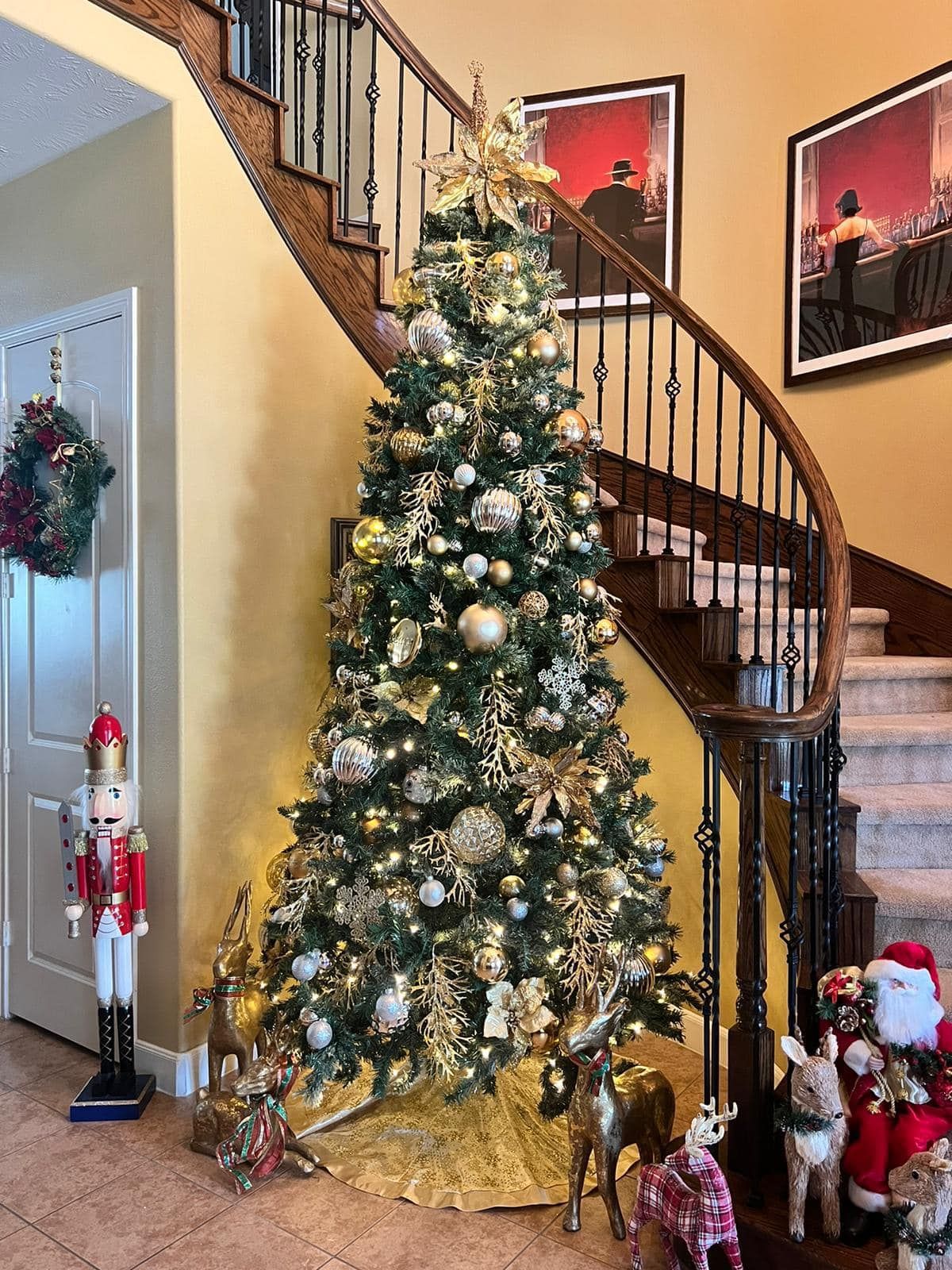 A tall, decorated Christmas tree stands in a foyer by a staircase, flanked by a nutcracker, reindeer, and Santa figures.